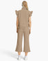 A model shown from the back wearing a beige ribbed knit two-piece set consisting of a sleeveless mock-neck top with ruffled shoulders and matching wide-leg cropped pants, paired with white sneakers against a light gray studio background.