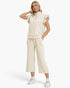 A woman modeling a matching cream-colored loungewear set consisting of a short-sleeve ruffled mock-neck zip sweatshirt and wide-leg cropped sweatpants, paired with white sneakers against a plain white background.