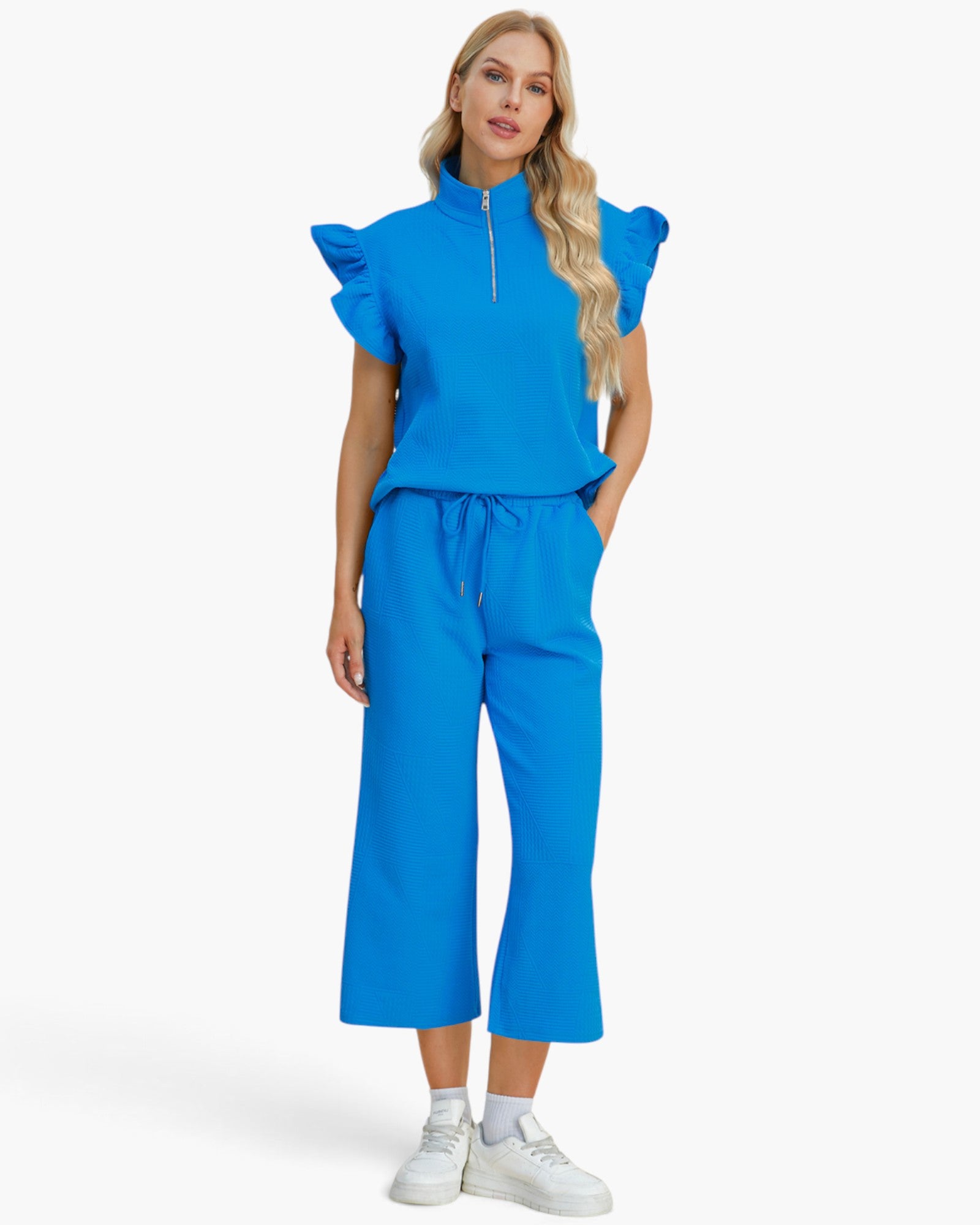 A woman modeling a bright blue two-piece outfit consisting of a short-sleeve top with ruffled shoulders and a half-zip neckline, paired with matching wide-leg cropped pants. She is wearing white sneakers against a clean white studio background.