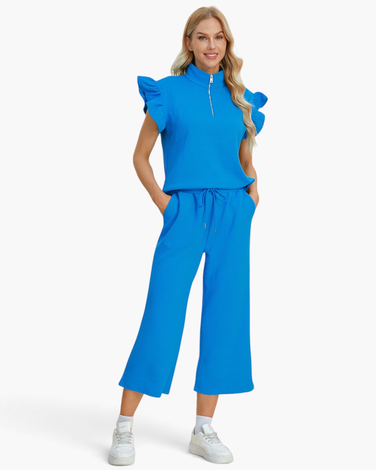 A woman modeling a bright blue two-piece outfit consisting of a ruffled-sleeve half-zip top and matching wide-leg cropped pants, paired with white sneakers against a light gray studio background.