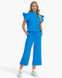 A woman modeling a bright blue two-piece outfit consisting of a ruffled-sleeve half-zip top and matching wide-leg cropped pants, paired with white sneakers against a light gray studio background.