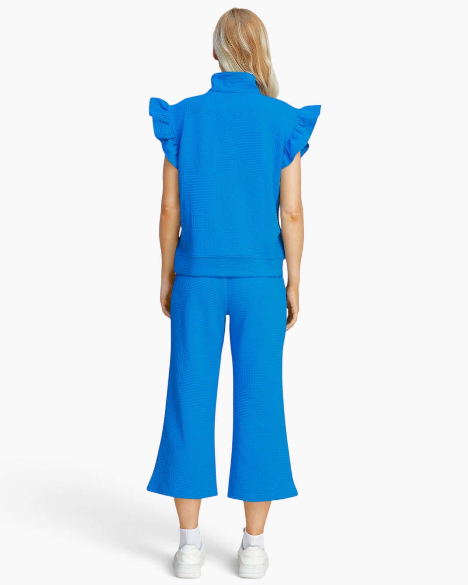 A model viewed from the back wearing a bright blue matching set consisting of a short-sleeve top with ruffled shoulders and high collar, paired with matching wide-leg cropped pants, on a light gray studio background.