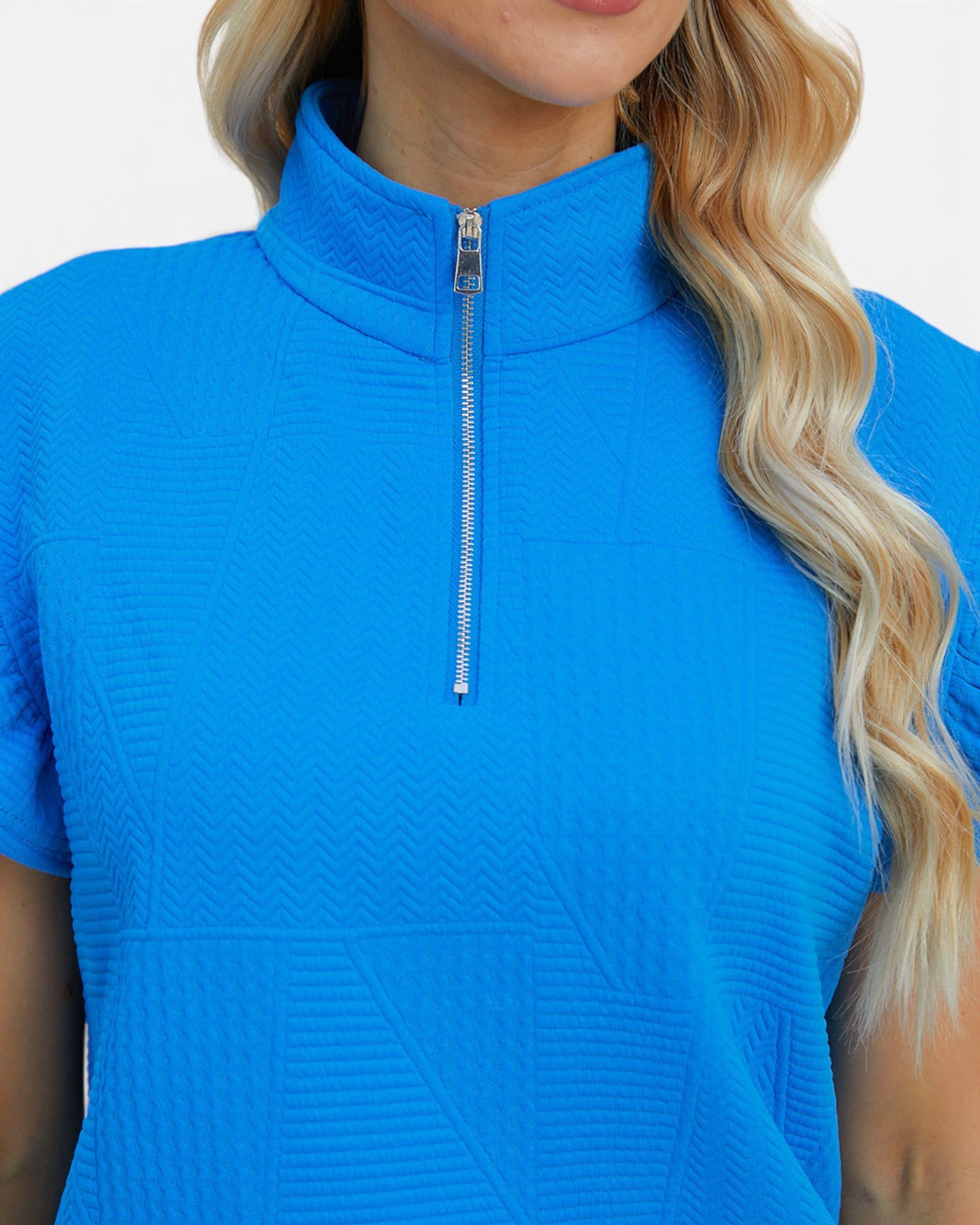 A close-up of a woman wearing a bright blue short-sleeve mock neck top with a silver zipper and textured geometric pattern.