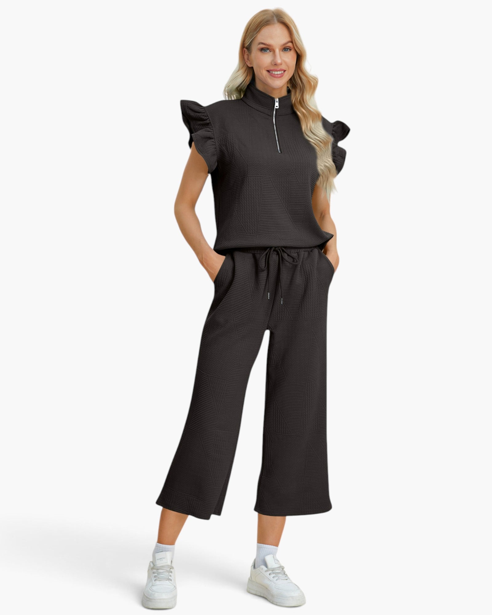 A woman modeling a dark charcoal gray two-piece outfit consisting of a ruffled sleeve mock-neck top with a half-zip and matching wide-leg cropped pants, paired with white sneakers against a light gray studio background.