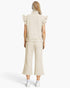 A model shown from the back wearing a cream-colored ribbed knit set consisting of a sleeveless mock-neck top with ruffled shoulders and matching cropped wide-leg pants, paired with white sneakers.