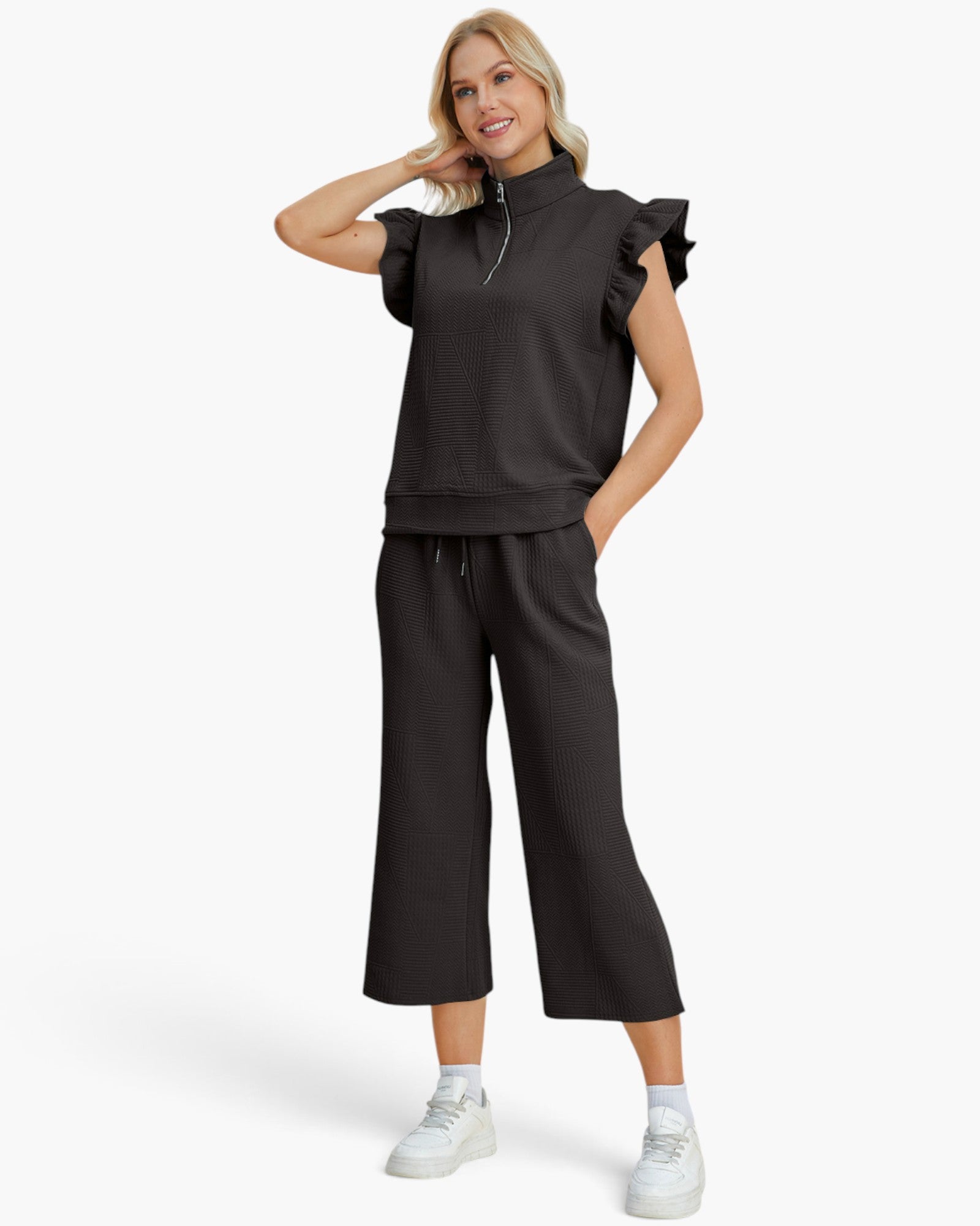 A woman modeling a black two-piece lounge set consisting of a short-sleeve mock-neck top with ruffled shoulder details and matching wide-leg cropped pants, paired with white sneakers against a light gray background.