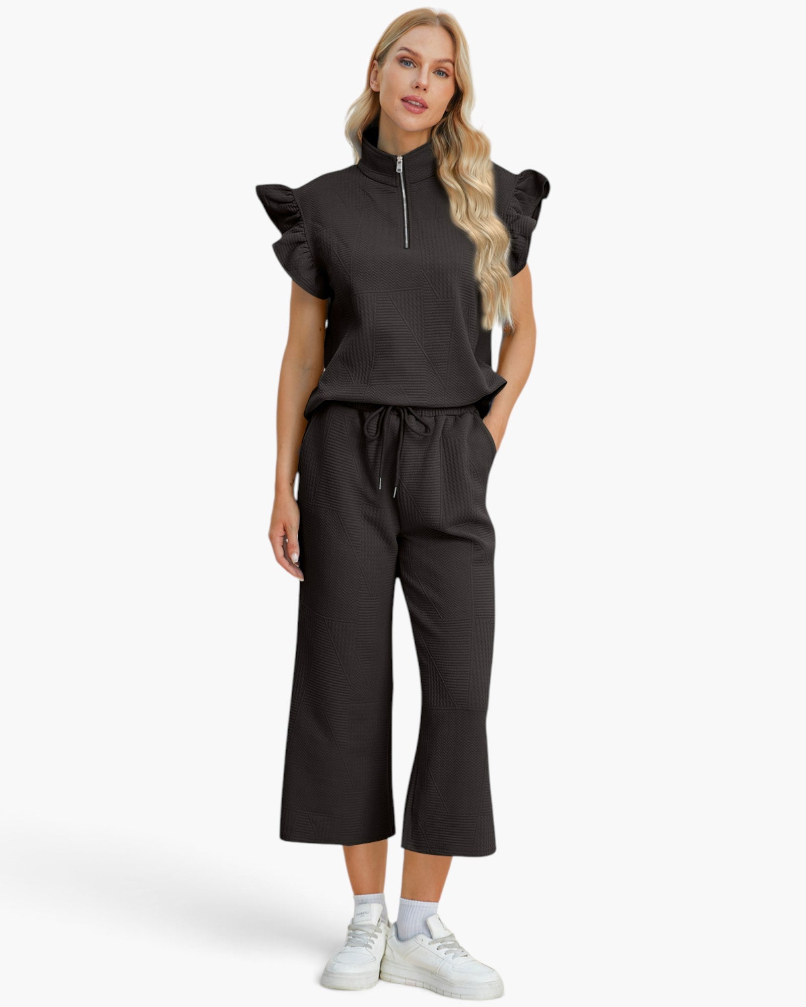 A woman modeling a black two-piece outfit consisting of a short-sleeve top with ruffled cap sleeves and a half-zip neckline, paired with matching black wide-leg cropped pants, against a plain light gray studio background.
