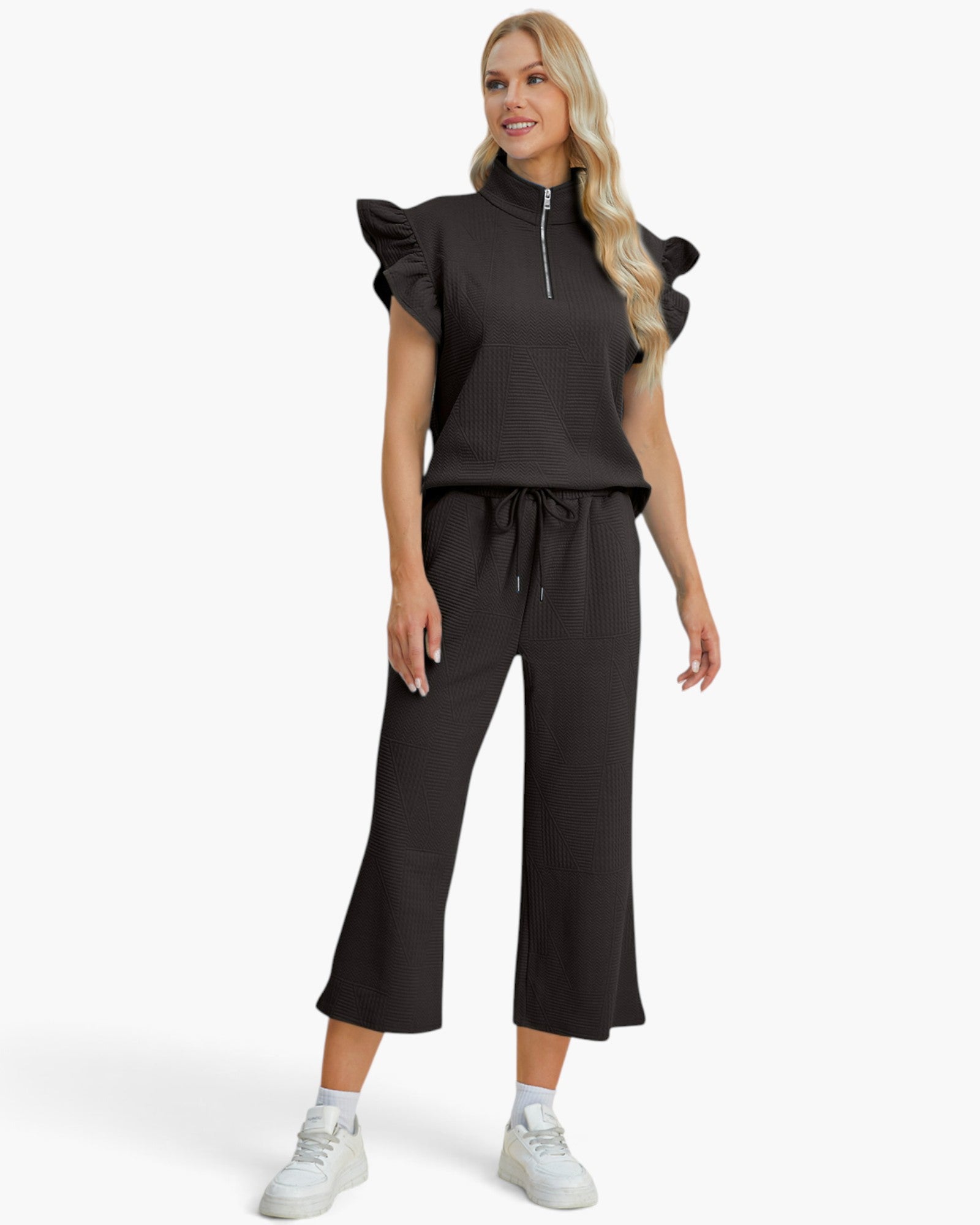 A woman modeling a black two-piece lounge set consisting of a short-sleeve top with ruffled shoulders and a half-zip neckline, paired with matching wide-leg cropped pants with a drawstring waist, on a light gray studio background.