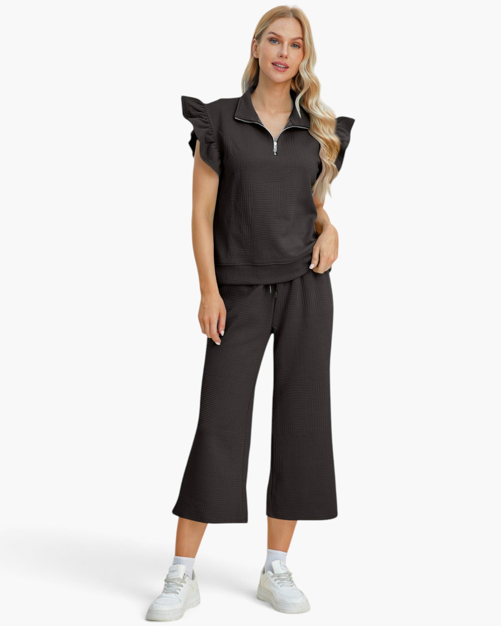A woman modeling a matching black two-piece lounge set consisting of a ruffle-sleeve half-zip sweatshirt and wide-leg cropped pants, paired with white sneakers against a light gray studio background.