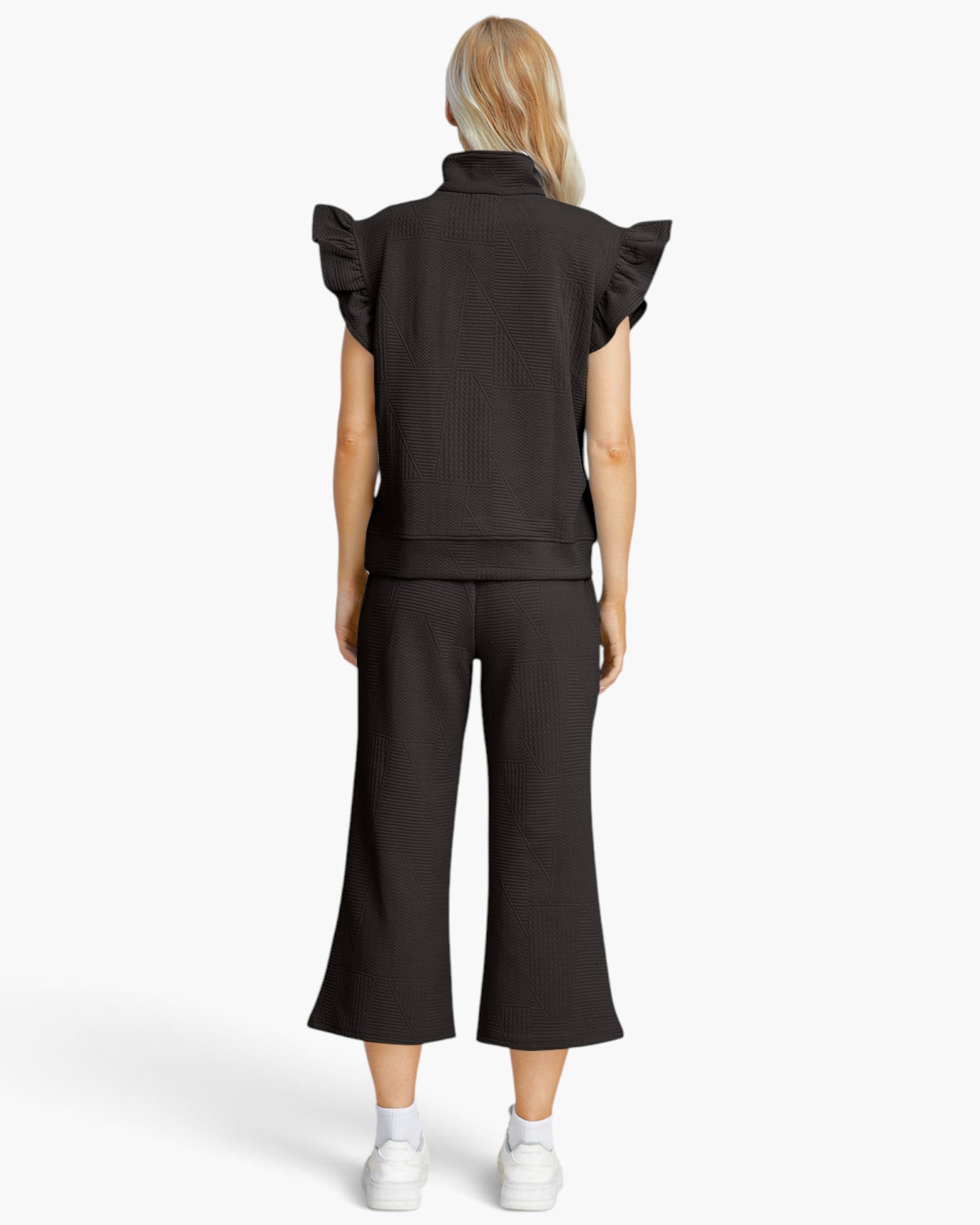 A model shown from the back wearing a black ribbed knit two-piece set consisting of a short-sleeve top with ruffled shoulders and matching wide-leg cropped pants, paired with white sneakers on a light gray background.
