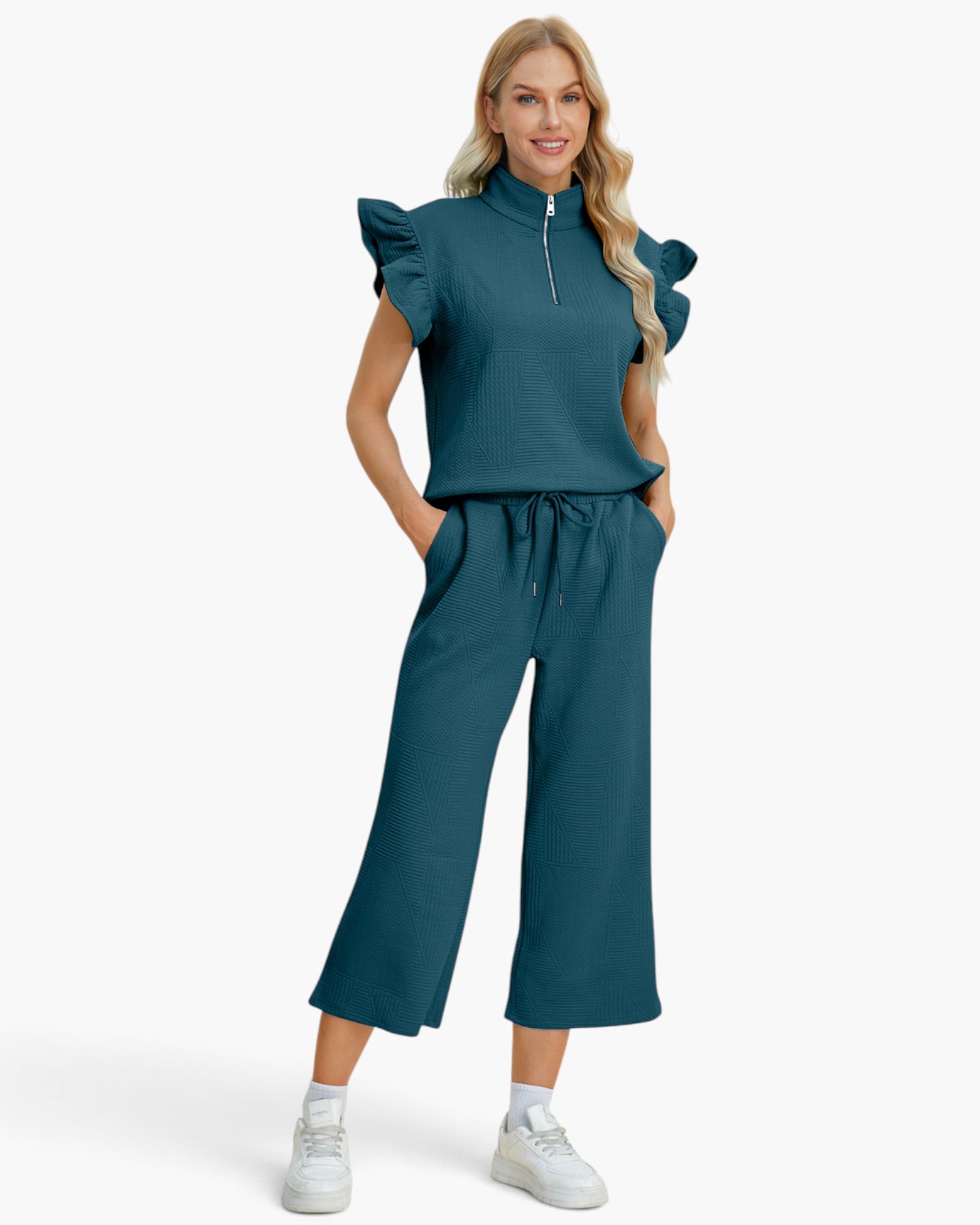 A woman modeling a matching two-piece outfit consisting of a teal ruffle-sleeve half-zip top and matching wide-leg cropped pants, paired with white sneakers against a light gray studio background.