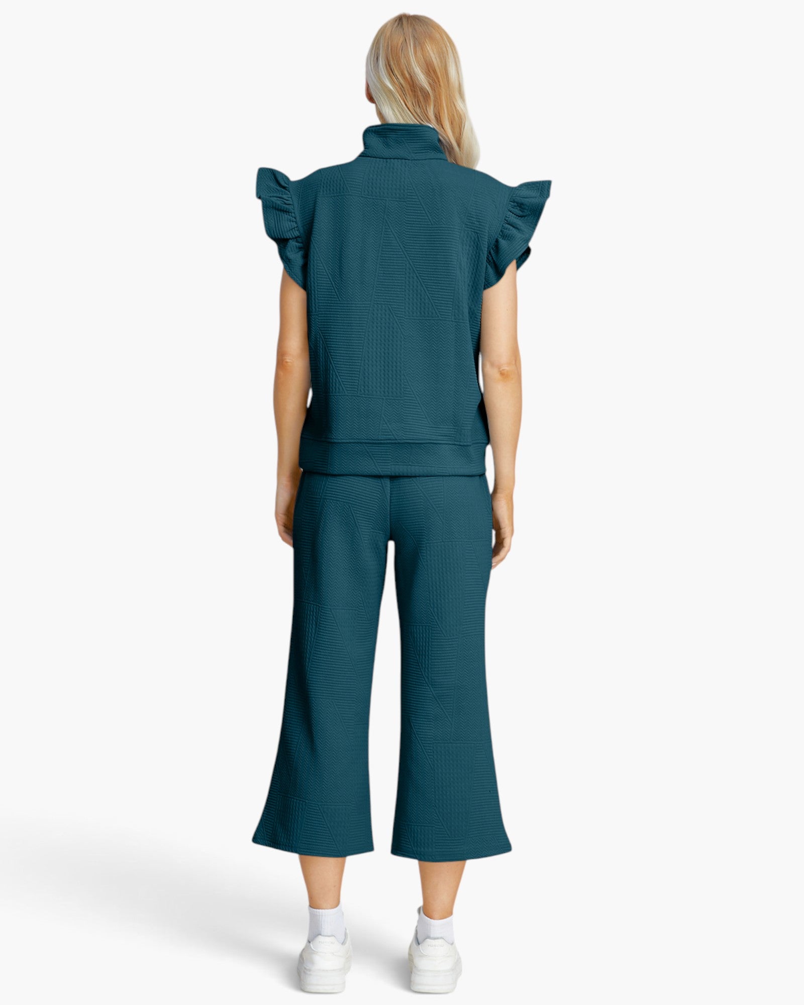 A model viewed from the back wearing a matching dark teal ribbed two-piece set consisting of a short-sleeve top with ruffled shoulders and wide-leg cropped pants, paired with white sneakers on a light gray background.