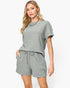 A woman modeling a light sage green crinkled two-piece lounge set consisting of a short-sleeve top and matching drawstring shorts, set against a plain white studio background.