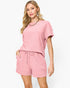A woman modeling a pink two-piece lounge set featuring a short-sleeve top and matching drawstring shorts, set against a plain white studio background.
