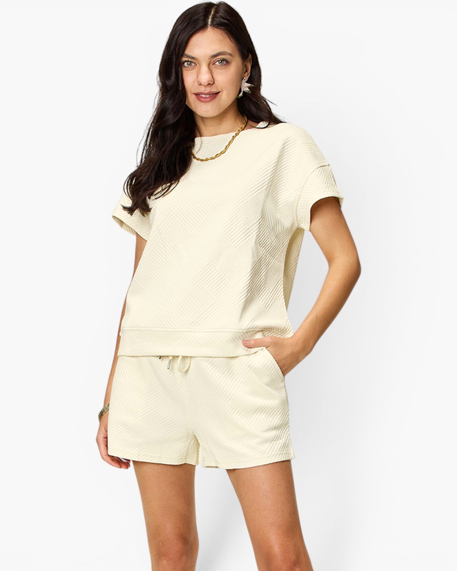 A woman modeling a matching cream-colored lounge set consisting of a short-sleeve textured top and coordinating shorts, set against a plain light gray background.
