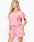 A woman modeling a pink textured short-sleeve top and matching drawstring shorts set against a plain white studio background.