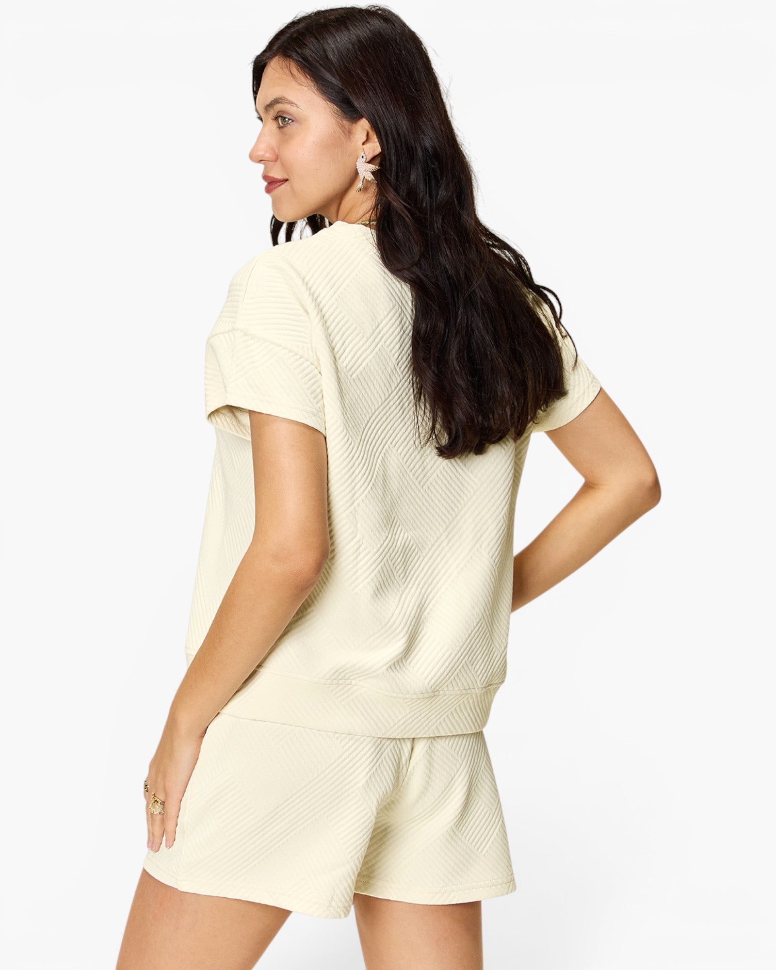 A woman wearing a cream-colored textured knit short-sleeve top and matching shorts, shown from a back-three-quarter angle against a plain white studio background.