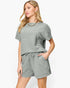 A woman modeling a matching two-piece lounge set in a textured sage green fabric, featuring a short-sleeve top and drawstring shorts, against a plain white background.