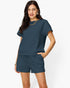 A woman modeling a matching two-piece lounge set consisting of a short-sleeve textured top and coordinating shorts in a muted slate blue color, set against a plain white studio background.