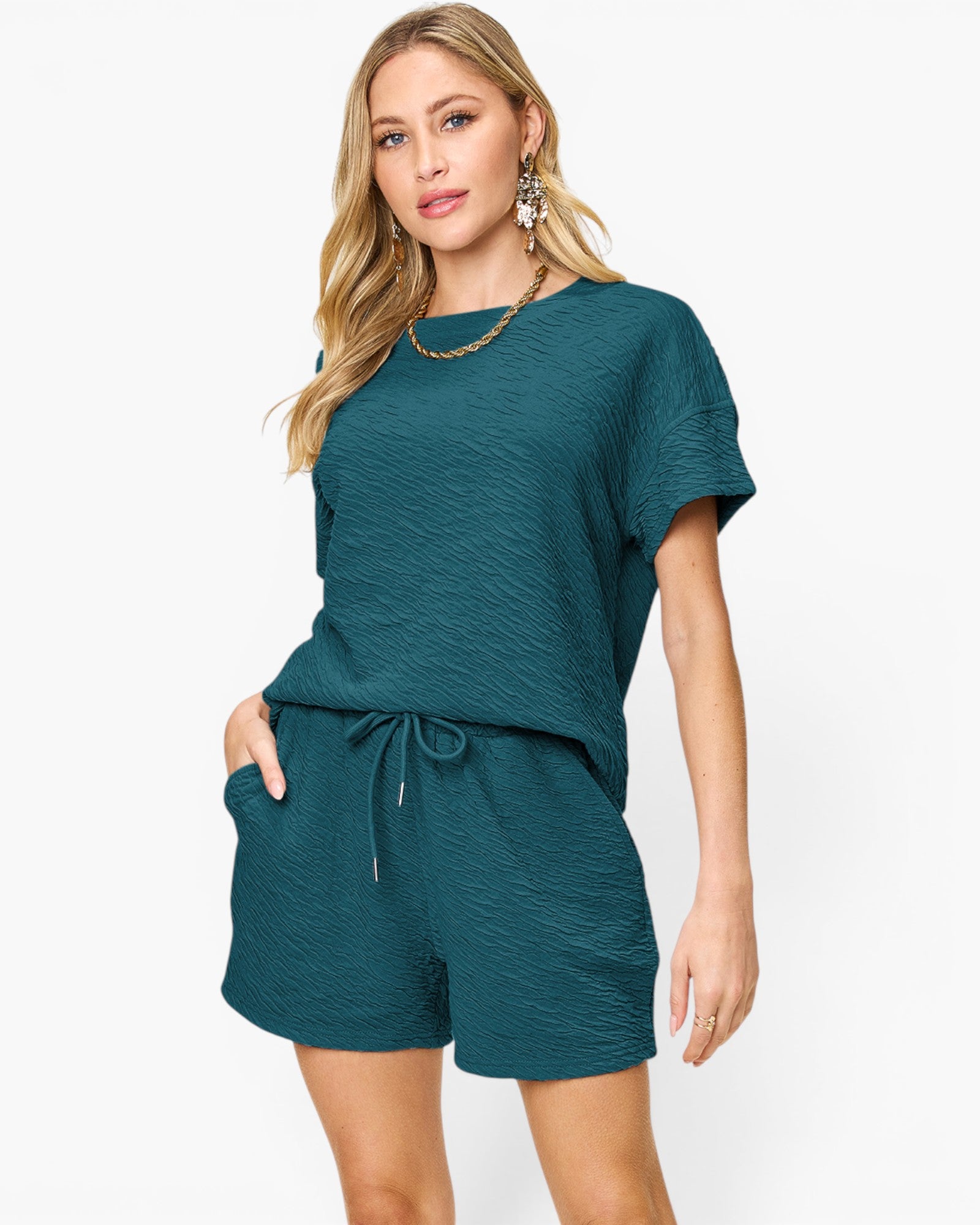 A woman modeling a matching two-piece lounge set in a textured teal green fabric, featuring a short-sleeve top and drawstring shorts, against a plain white background.