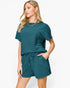 A woman modeling a matching teal short-sleeve top and shorts set with a crinkled texture, shown against a plain light gray studio background.