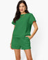 A woman modeling a matching green lounge set consisting of a short-sleeve top and drawstring shorts, photographed against a clean white studio background.