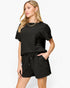 A woman modeling a black textured short-sleeve top and matching black drawstring shorts against a light gray studio background.