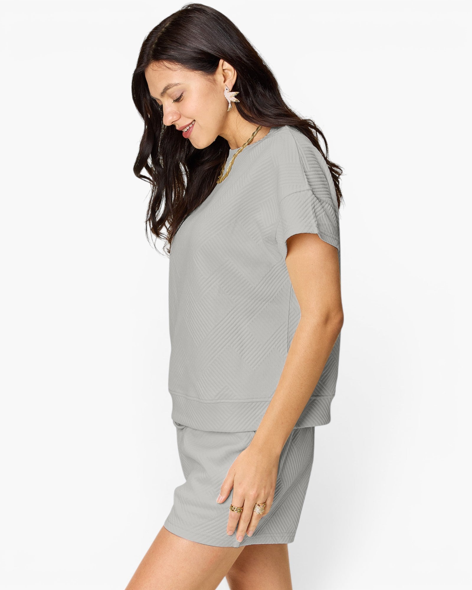 A woman wearing a light gray knit two-piece set consisting of a short-sleeve top and matching mini skirt, shown from a side profile against a plain white studio background.