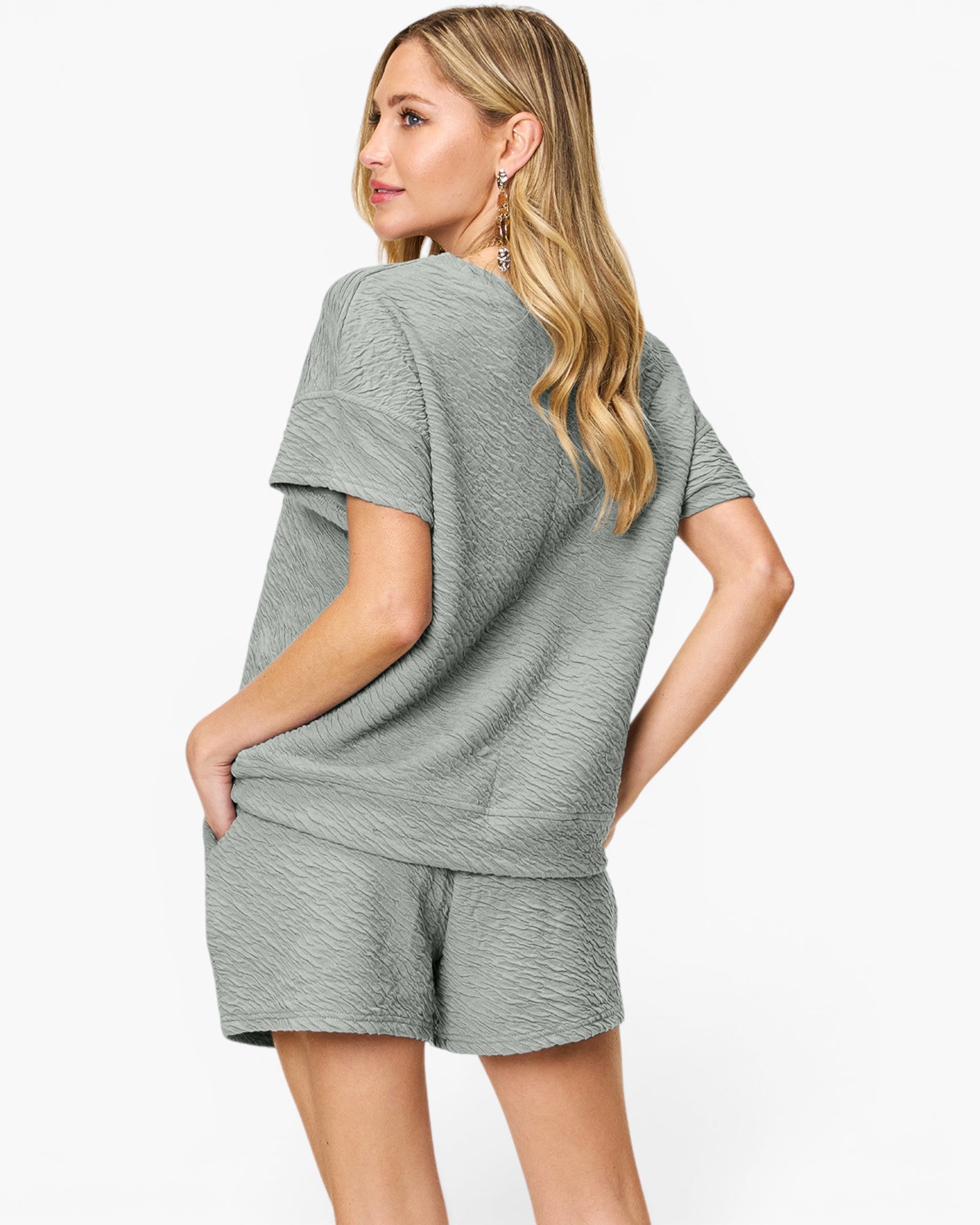 A woman modeling a matching two-piece lounge set consisting of a short-sleeve top and shorts in a textured sage green fabric, shown from a three-quarters back view against a light gray studio background.