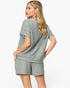 A woman modeling a matching two-piece lounge set consisting of a short-sleeve top and shorts in a textured sage green fabric, shown from a three-quarters back view against a light gray studio background.