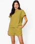 A woman modeling a matching two-piece lounge set consisting of a short-sleeve textured top and drawstring shorts in a muted olive-mustard yellow color, set against a plain light gray background.