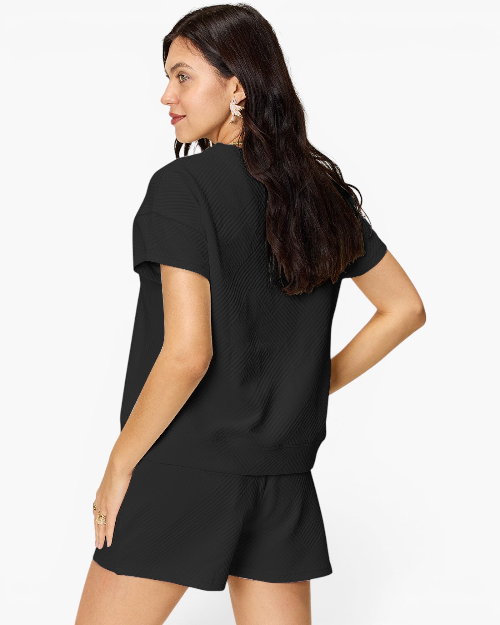 A woman wearing a black short-sleeve textured top and matching black shorts, shown from a side-back angle against a plain white studio background.