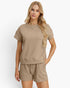 A woman modeling a matching beige knit lounge set consisting of a short-sleeve crewneck top and drawstring shorts, against a clean white background.