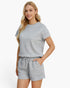 A woman modeling a light gray short-sleeve knit top and matching drawstring shorts against a plain white studio background.