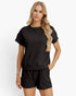 A blonde model wearing a matching black short-sleeve textured top and shorts set, standing against a clean white studio background.