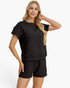 A woman modeling a matching black short-sleeve top and shorts set with a textured waffle-weave pattern, photographed against a clean white studio background.