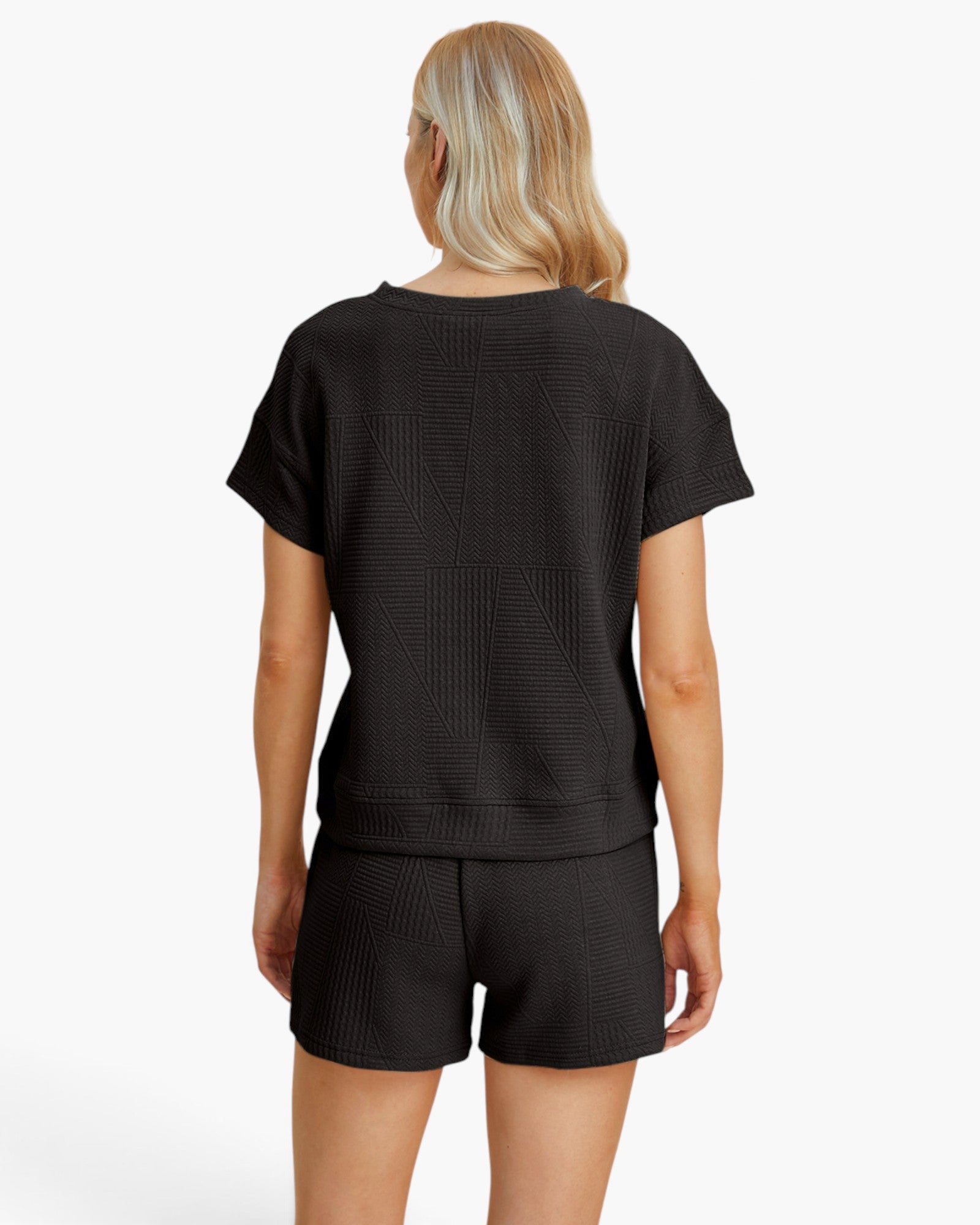 A woman shown from the back wearing a black short-sleeve cropped top and matching black shorts with a subtle geometric waffle texture, against a plain white studio background.