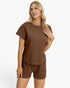 A woman modeling a matching brown lounge set consisting of a short-sleeve textured top and coordinating shorts against a plain white studio background.