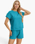 A woman modeling a matching two-piece lounge set in a vibrant teal color, featuring a short-sleeve textured top and coordinating shorts, set against a plain light gray background.