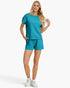 A woman modeling a matching turquoise blue lounge set consisting of a short-sleeve top and shorts, paired with white sneakers, against a plain white studio background.