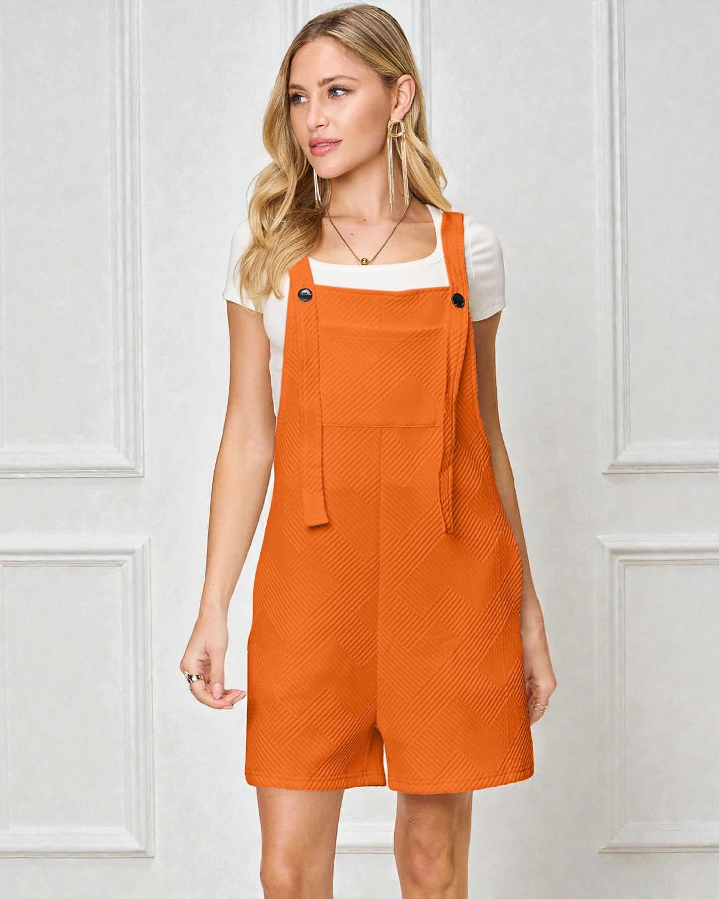 Woman wearing an orange denim pinafore dress against a white paneled wall.