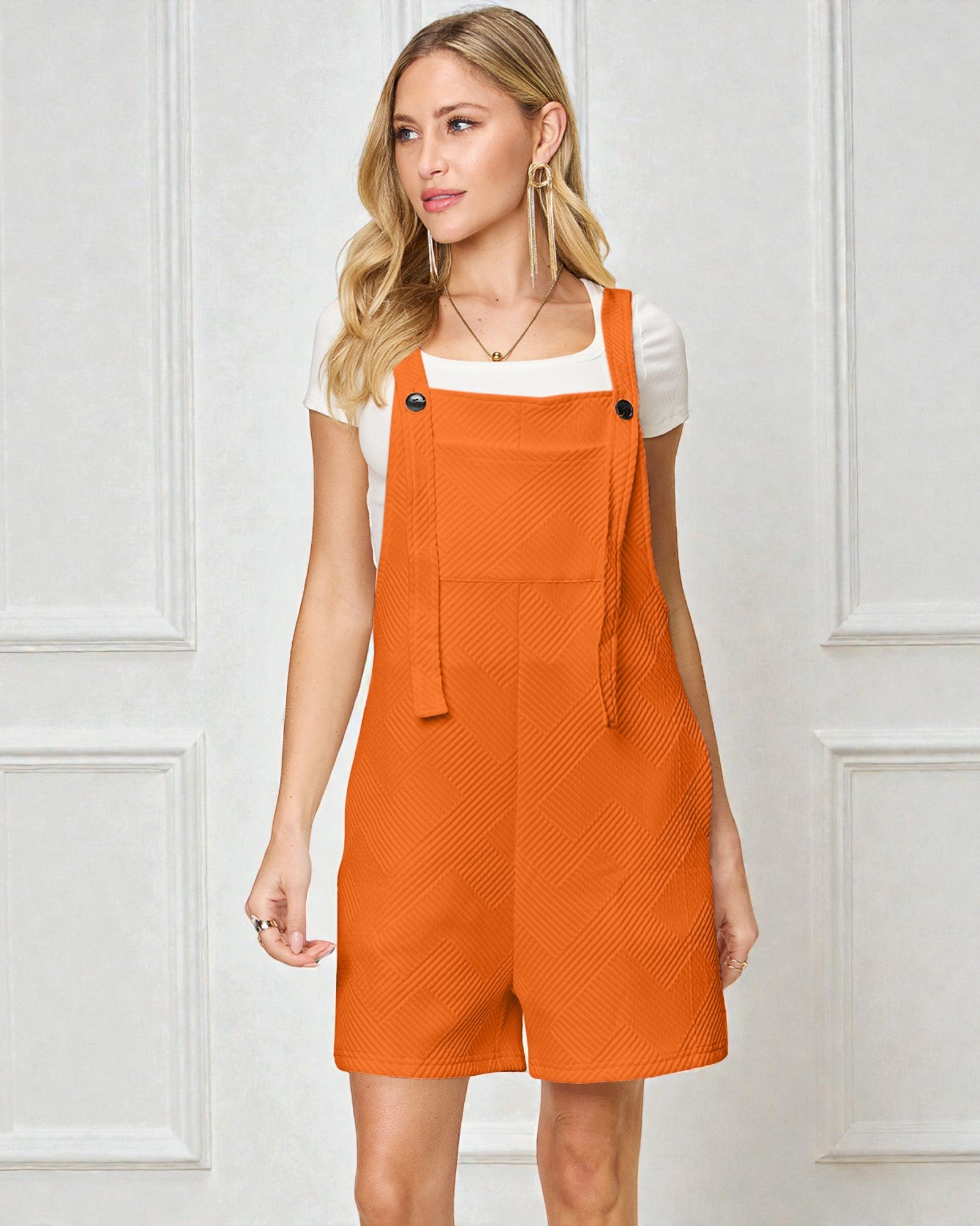 Woman wearing an orange denim pinafore dress against a white paneled wall.