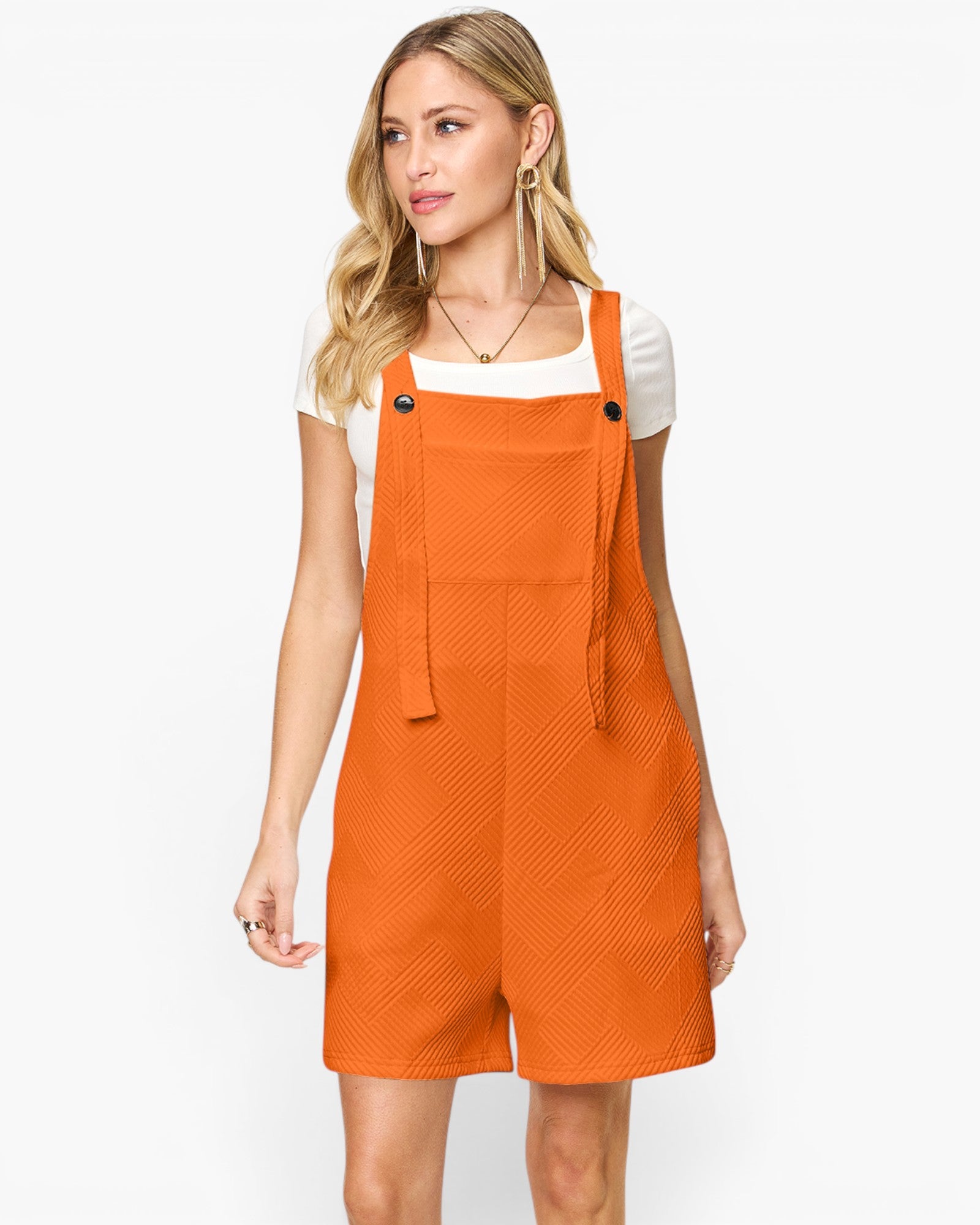 A woman modeling a bright orange textured overall romper with shoulder straps and black snap buttons, worn over a white t-shirt, against a plain white studio background.
