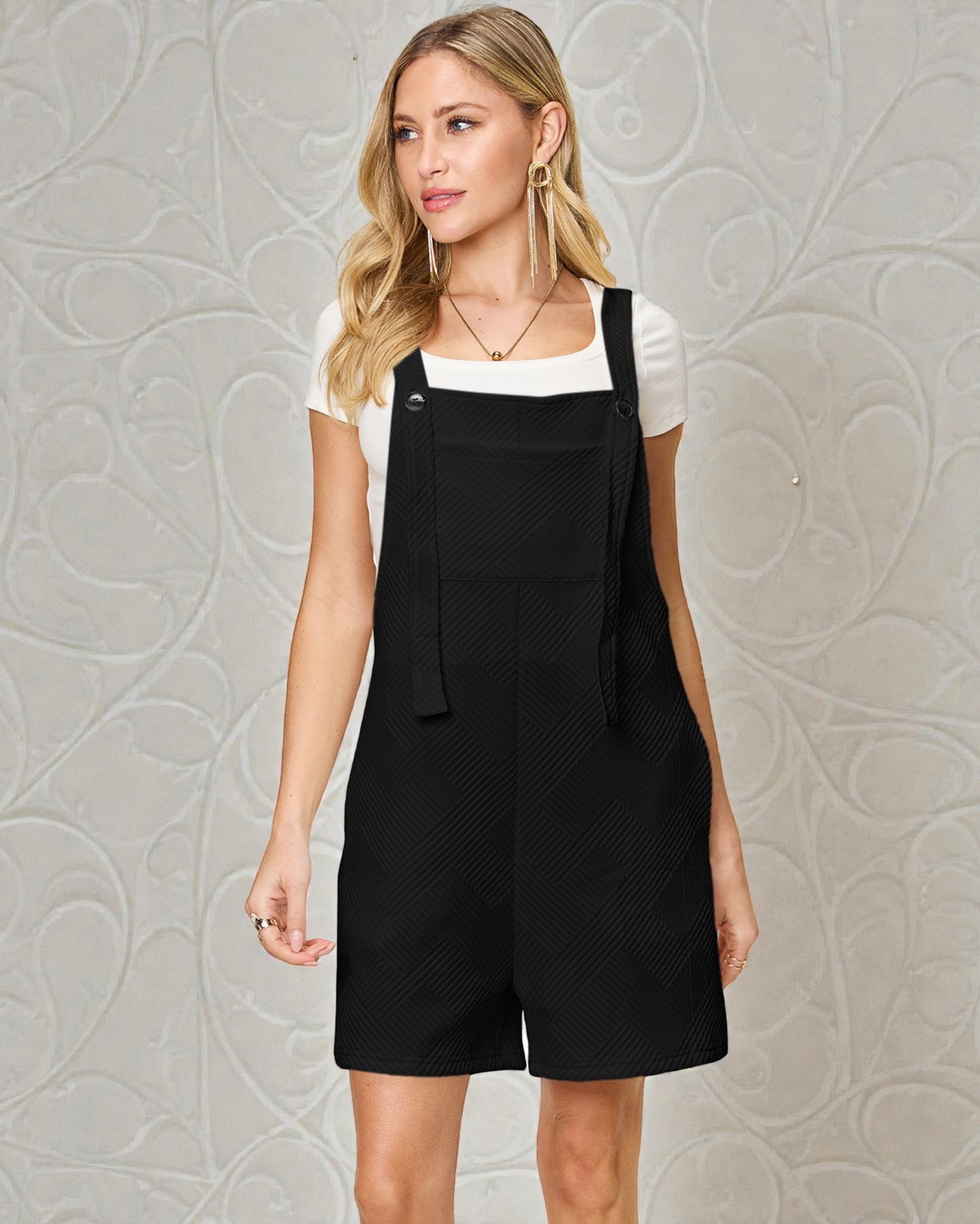 Woman wearing a black pinafore dress over a white shirt against a gray textured wall.