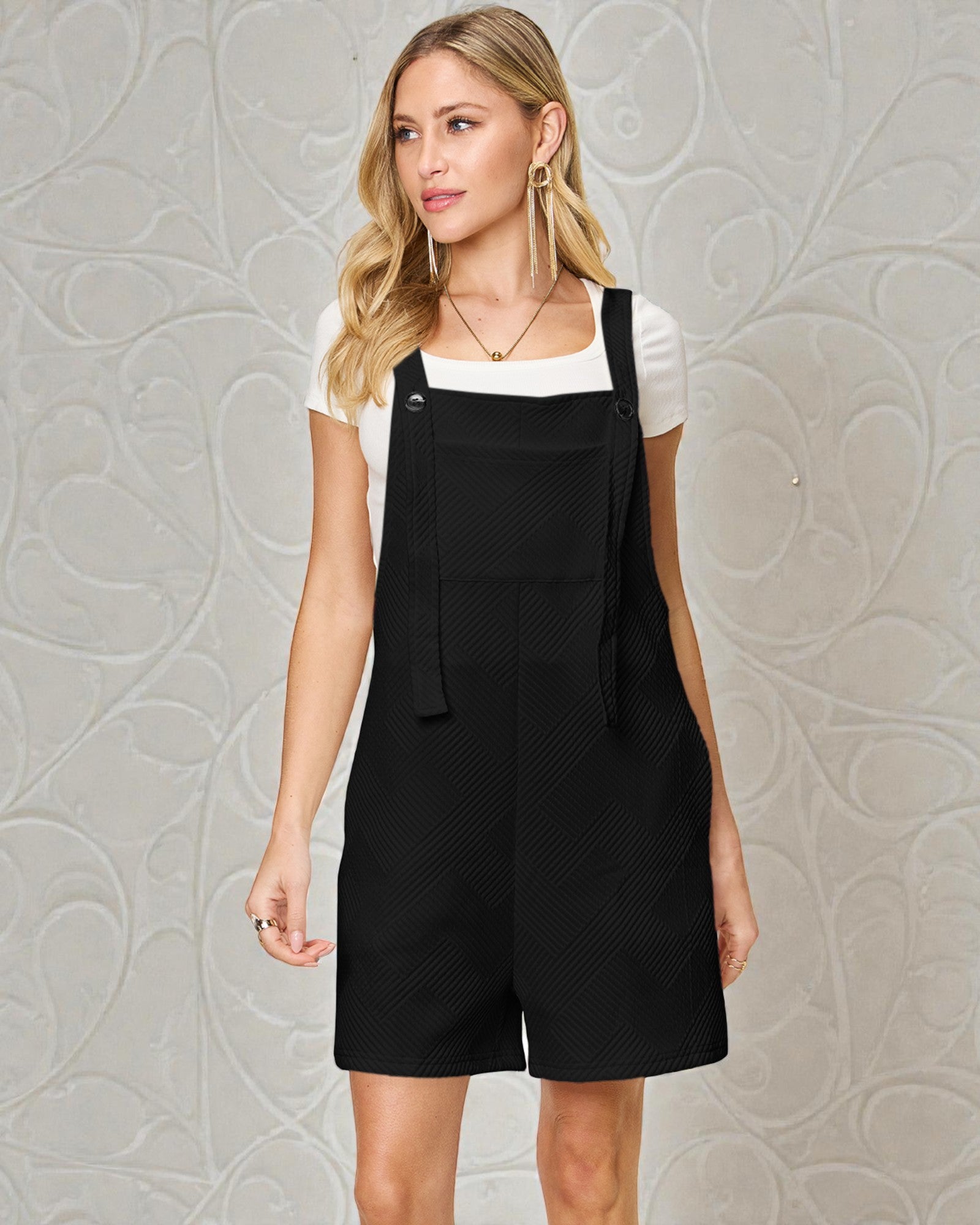 Woman wearing a black pinafore dress over a white shirt against a gray textured wall.