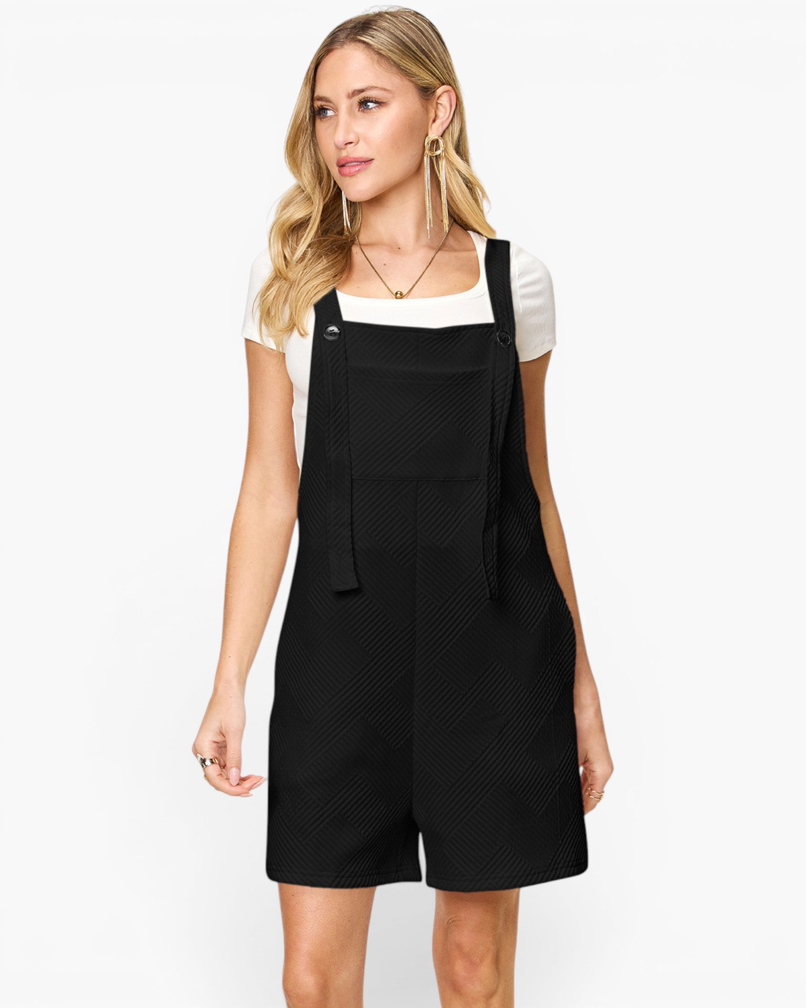 A woman modeling a black overall romper with a subtle textured pattern, worn over a white short-sleeve top, against a plain white studio background.
