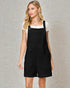Woman wearing a black pinafore dress over a white shirt against a gray textured wall.