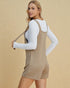 Woman wearing a beige pinafore dress over a white long-sleeve shirt against a beige background