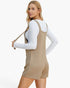 A woman modeling a beige textured overall romper with a white long-sleeve shirt underneath, holding a matching beige shoulder bag against a plain white studio background.