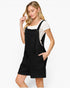 A woman modeling a black overall romper dress over a white short-sleeve t-shirt, photographed against a clean white studio background.